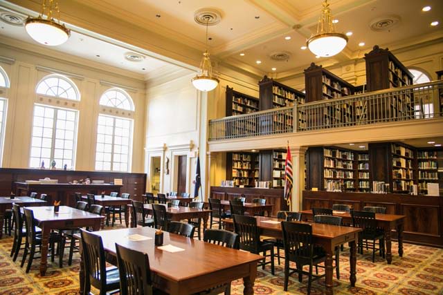 image of a large, two story Colonial Revival reading room. The ceiling and walls are cream colored. The shelves are filled with books and dark wood, which matches the tables.