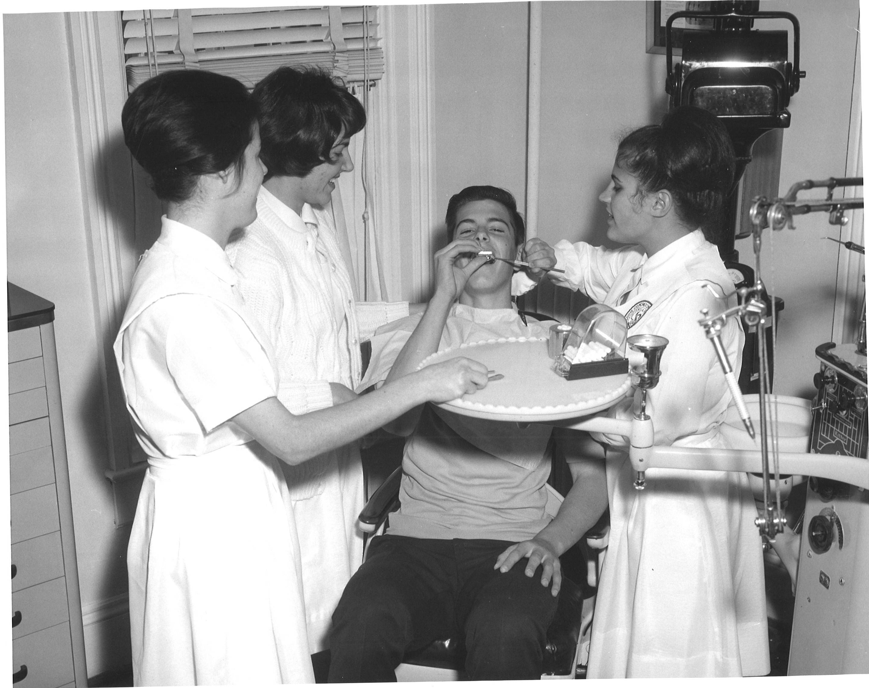 This image is of three white women dental hygiene students cleaning the teeth of a white male. One is seated as the other two pin on her hat.