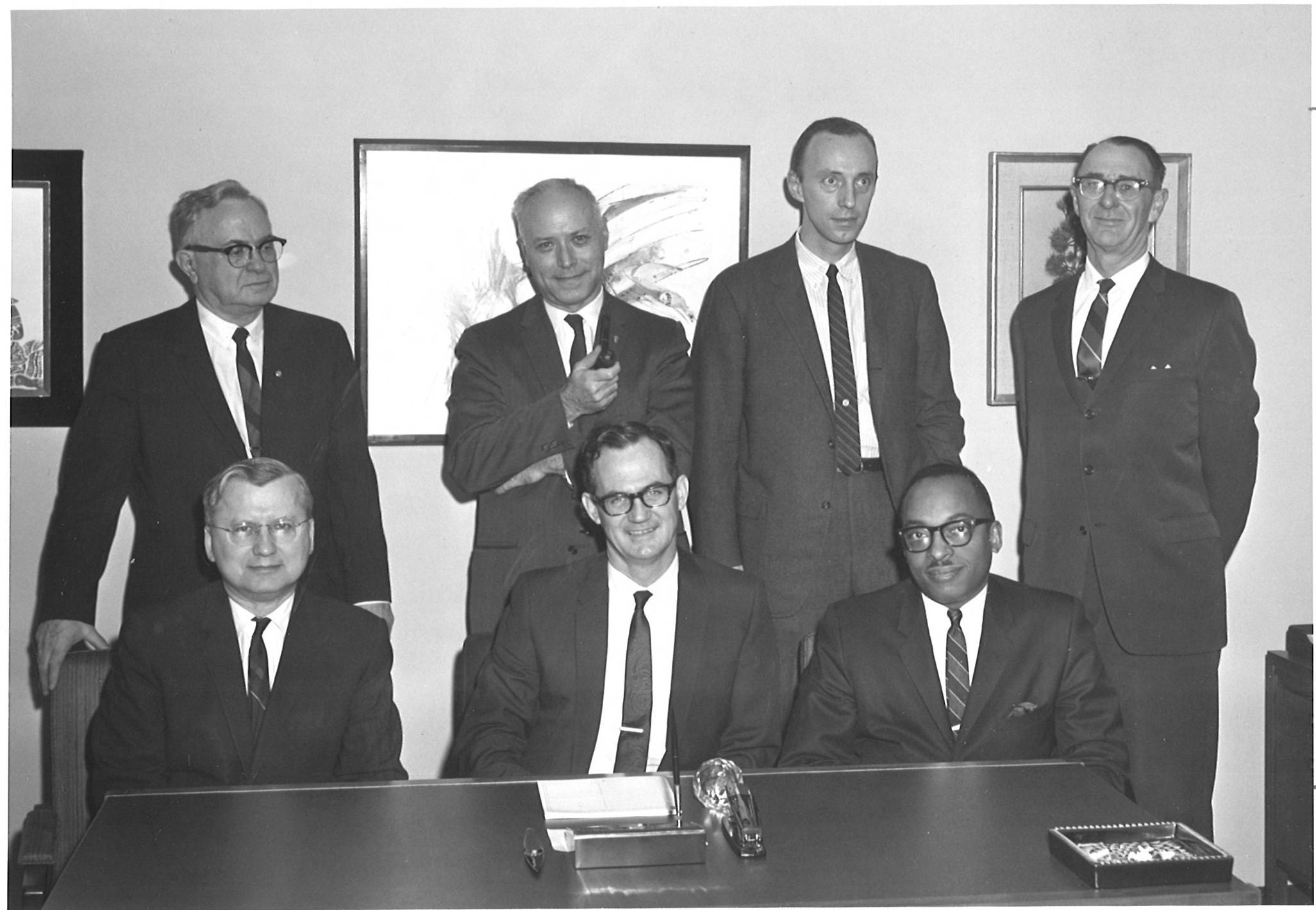This is a black and white image of seven men in suits. Four are standing in the back and three are sitting.