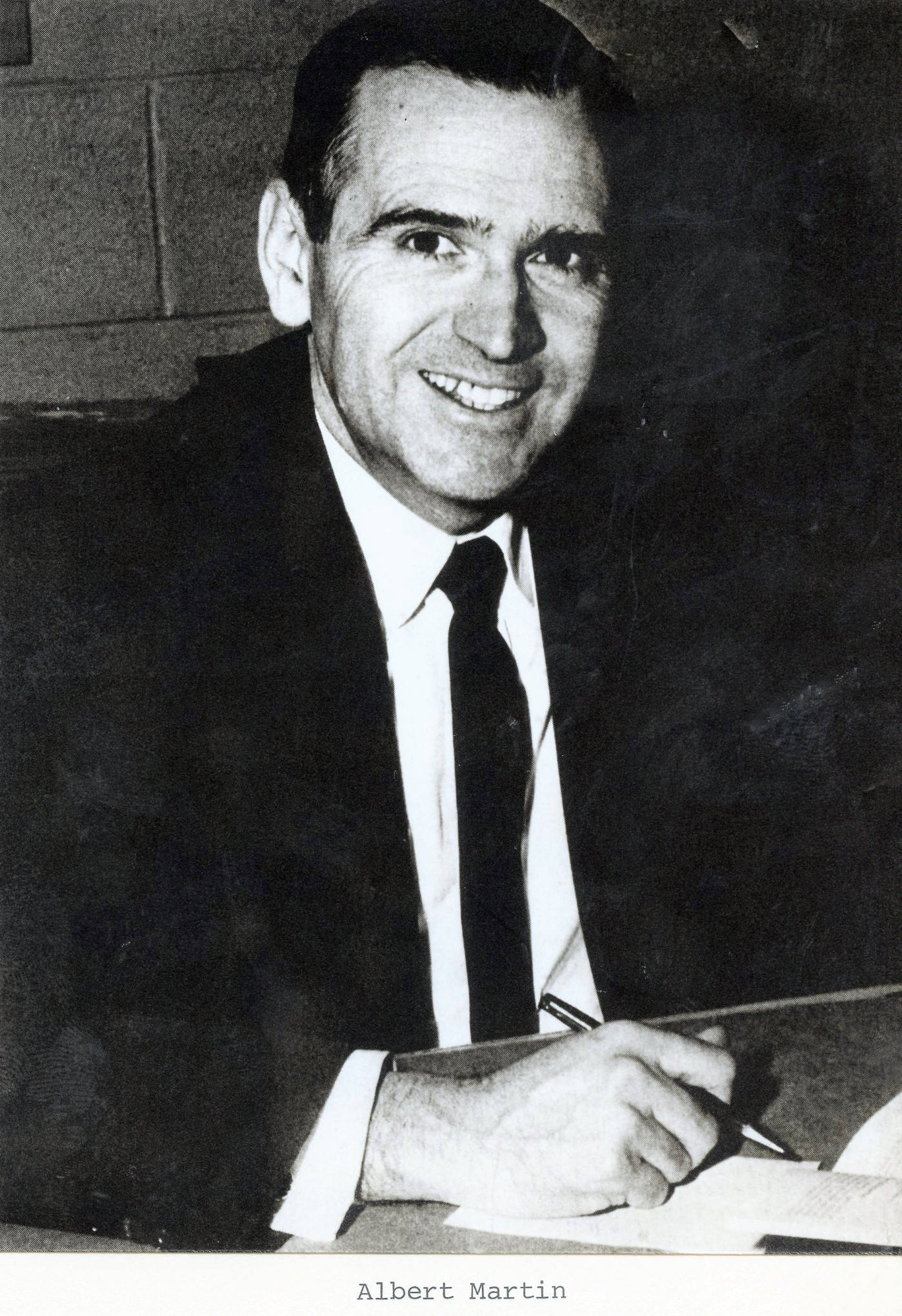 This is a black and white image is of a middle aged white man with dark hair and a smile. He is wearing a suit.