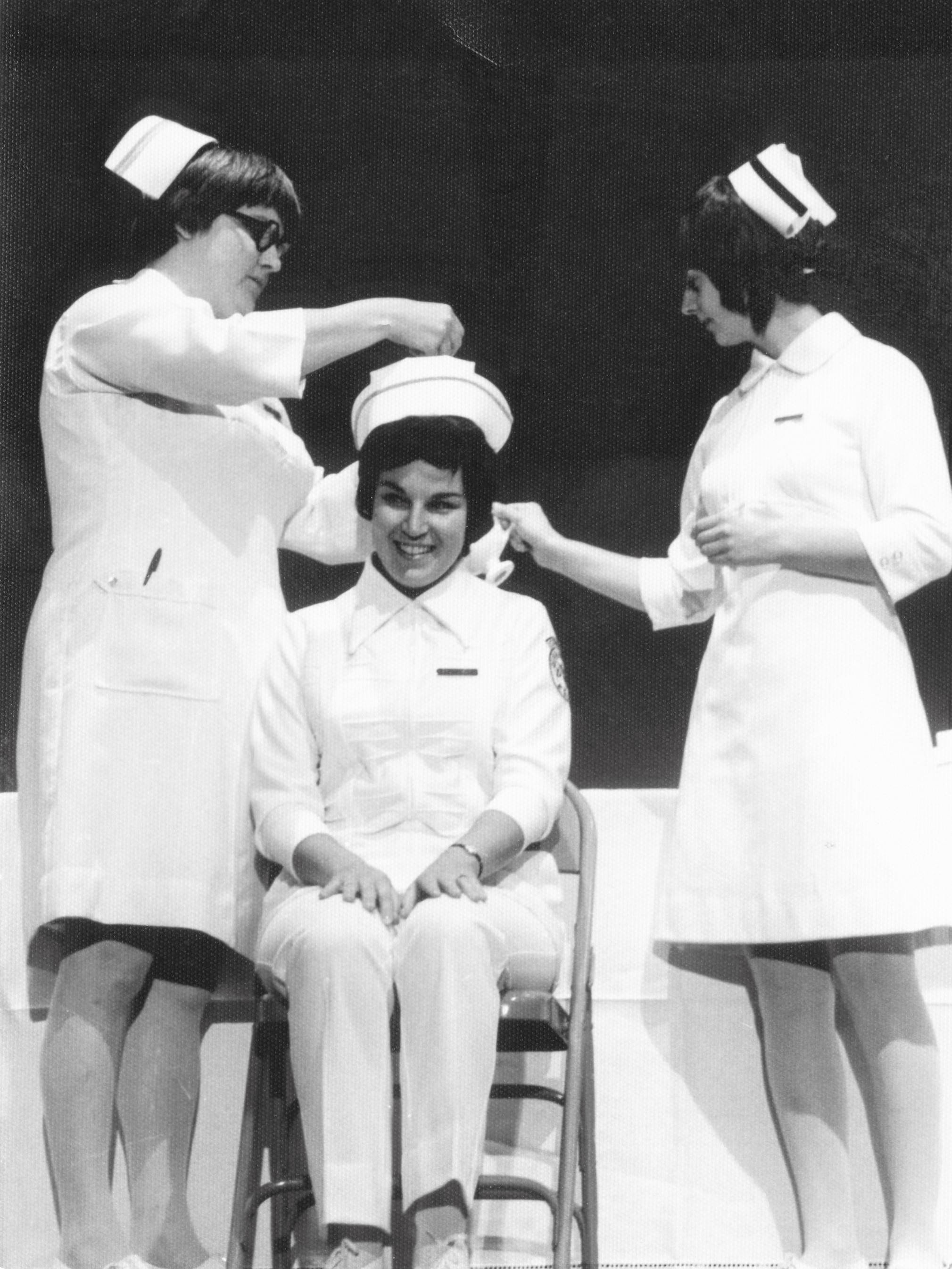 This image is of three white women in old fashioned nursing uniforms. One is seated as the other two pin on her cap.